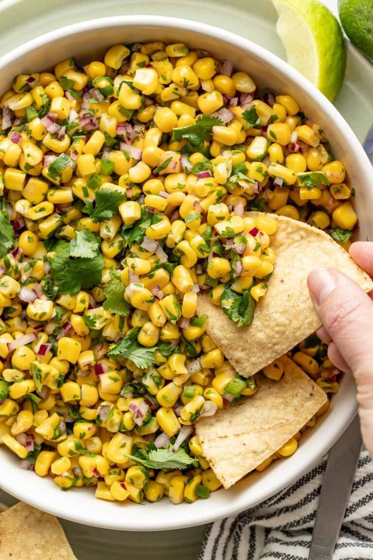 Overhead view of a bowl of Chipotle Corn Salsa with a hand scooping a tortilla chip into it.