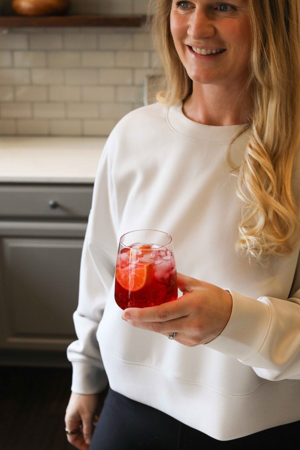 Woman in a kitchen holding a vibrant red mocktail with sliced fruit in the glass.