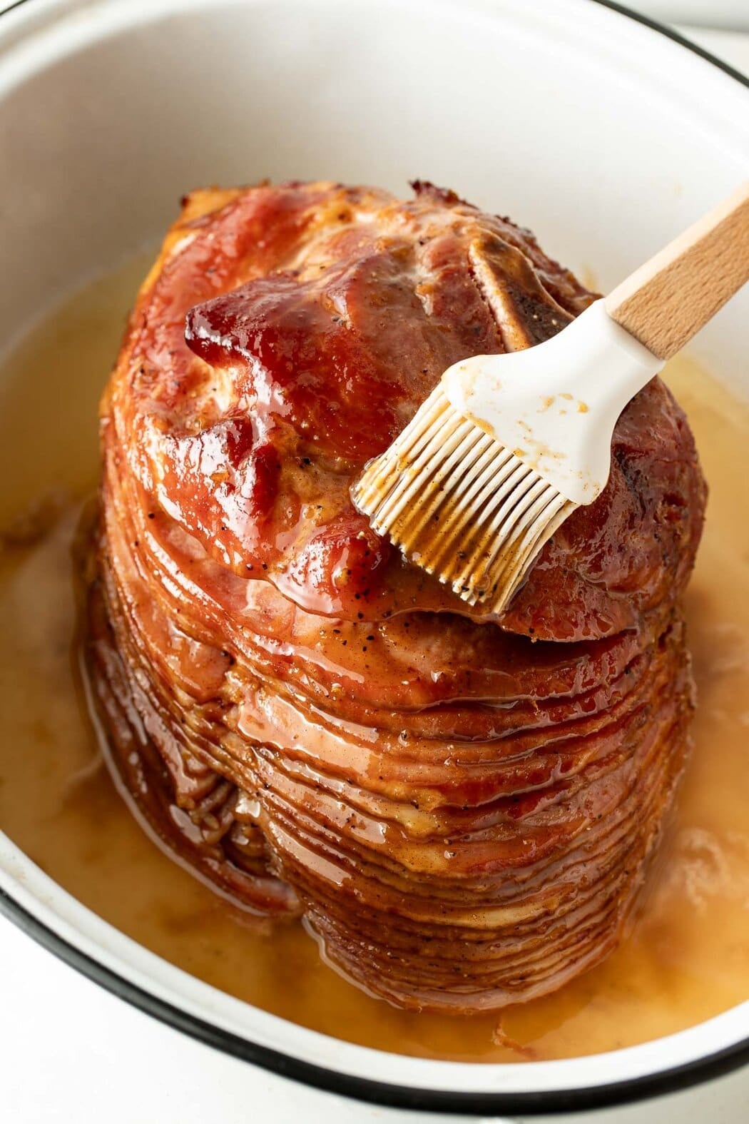 Close up view of a large ham in a roasting pan being brushed with a silicone brush that has ham glaze.