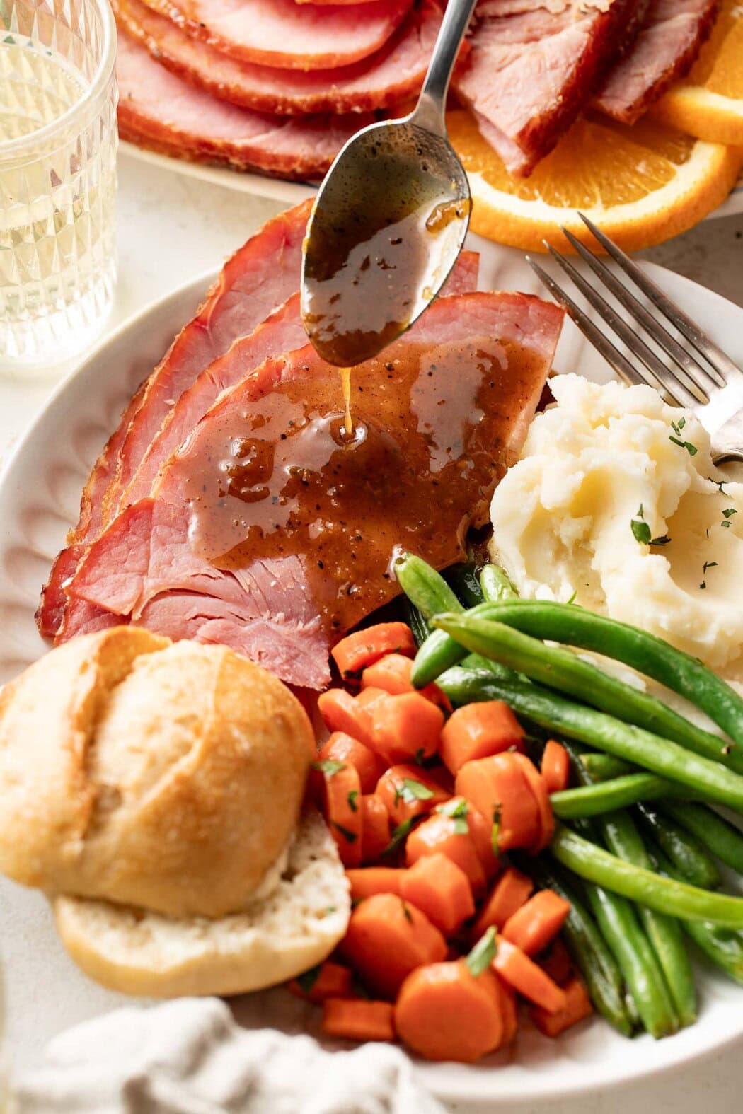 Overhead view of a plate of food with ham slices, mashed potatoes, green beans, roasted carrots, and a bun with a spoon drizzling ham glaze over the ham slices.