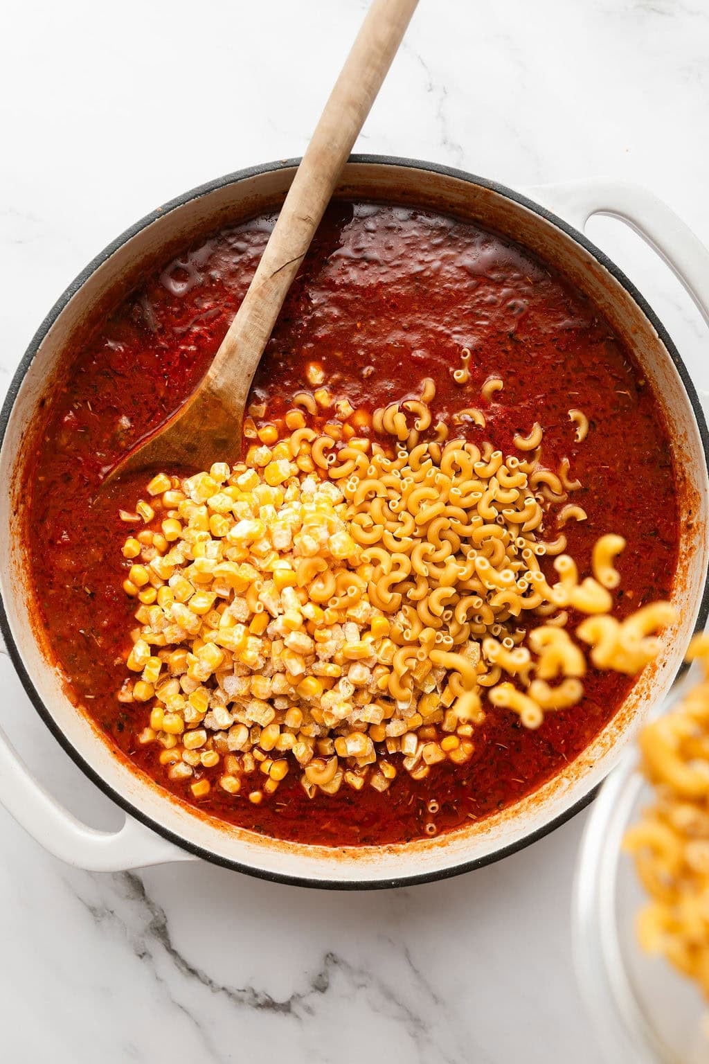Overhead view of a skillet filled with a tomato sauce and elbow macaroni being poured into the mixture.