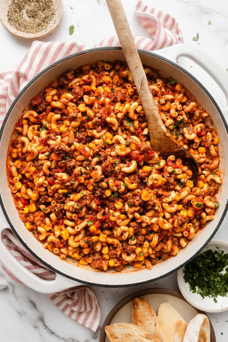 Overhead view of a white skillet filled with cooked goulash made with elbow macaroni and ground beef with a wooden serving spoon in the skillet.