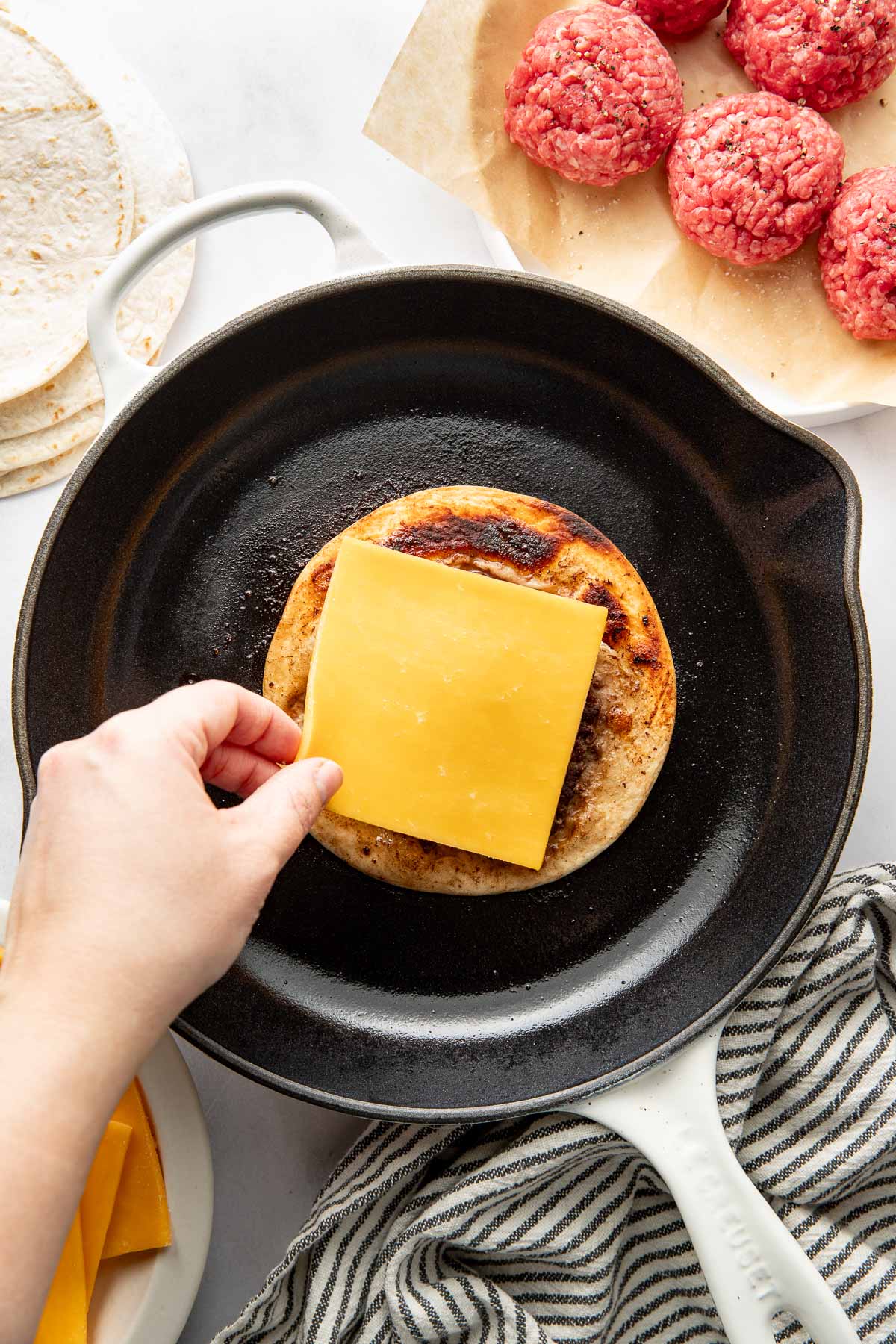 Overhead view of a fry pan with a Smash Burger Taco in it and a hand placing a piece of cheddar cheese on top.