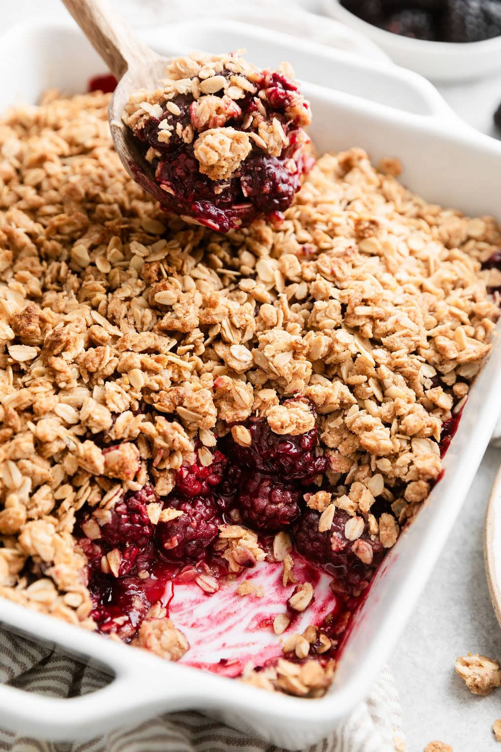 Close up view of a baking dish filled with freshly baked Blackberry Crumble with a wooden spoon bringing out a spoonful.