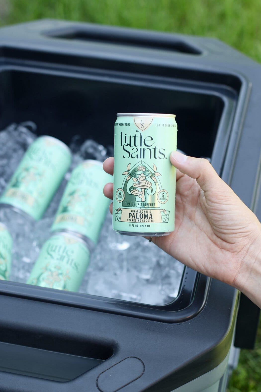 Close up view of a hand holding a little saints canned mocktail over a cooler filled with ice.