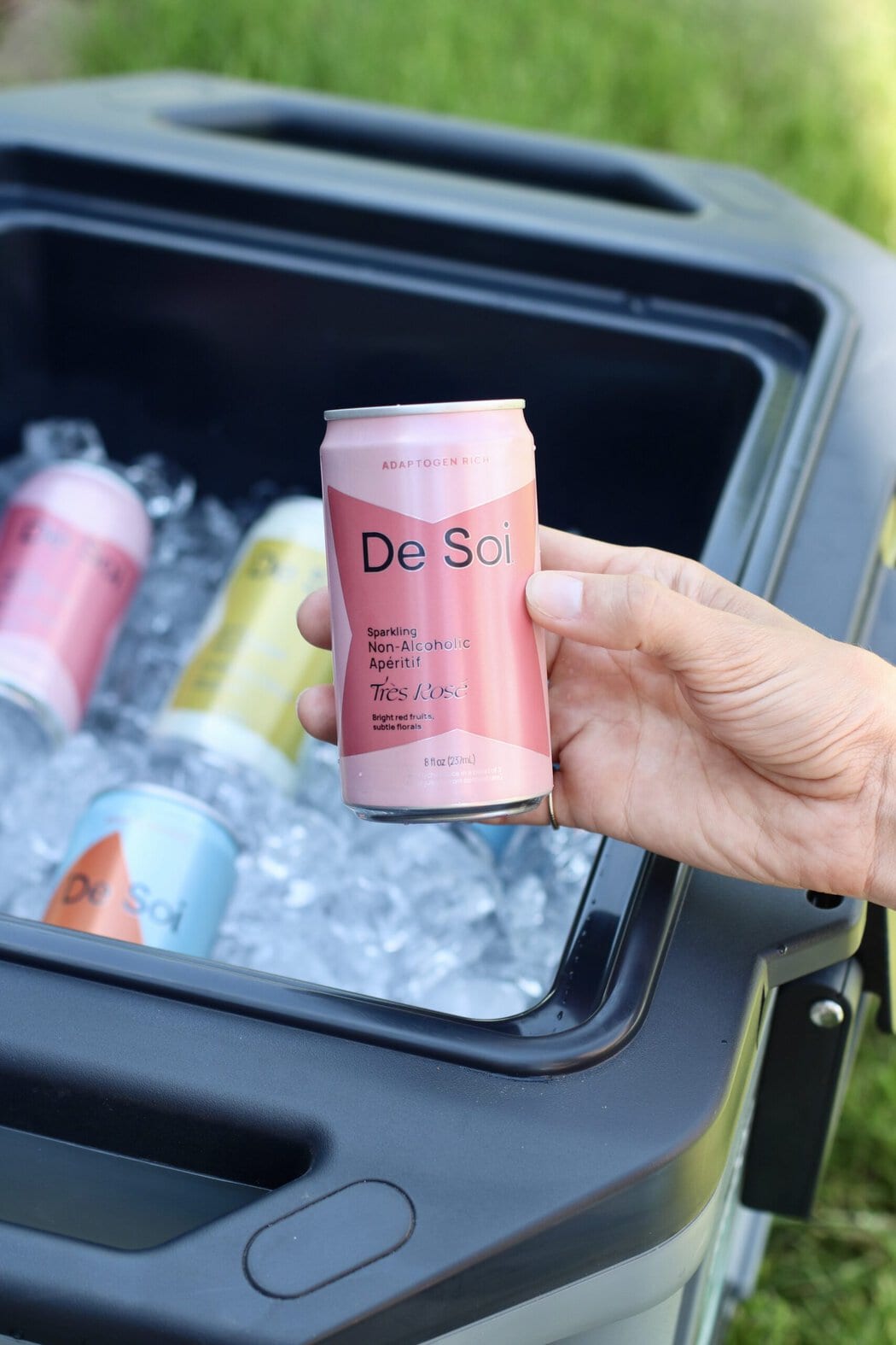 Close up view of a hand holding a pink de soi canned mocktail over a cooler full of ice.