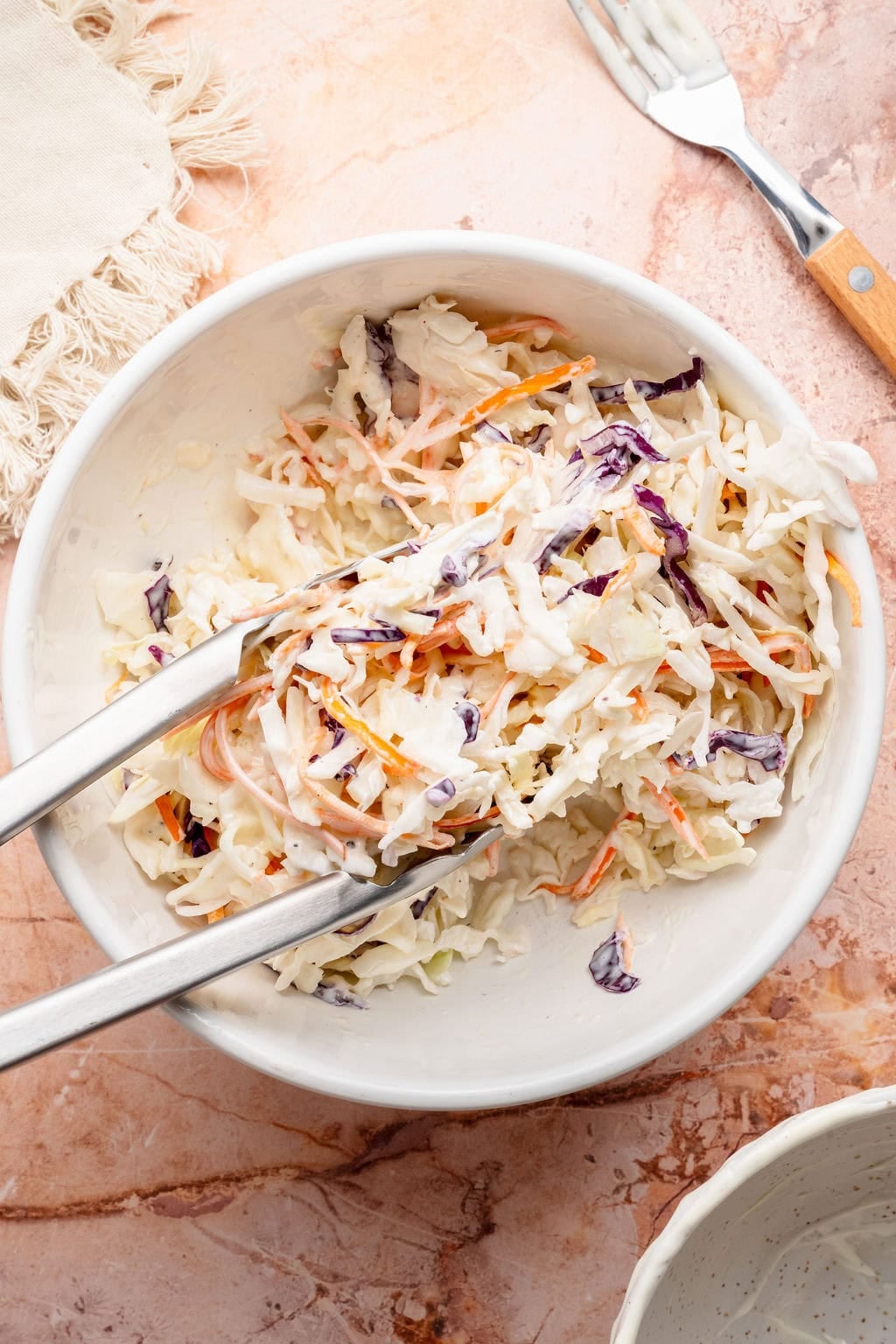 Overhead view of a stainless-steel tongs stirring a bowl of coleslaw.