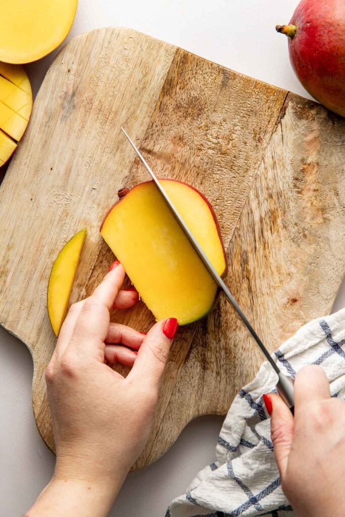 Overhead view of a mango being sliced on a wooden cutting board.