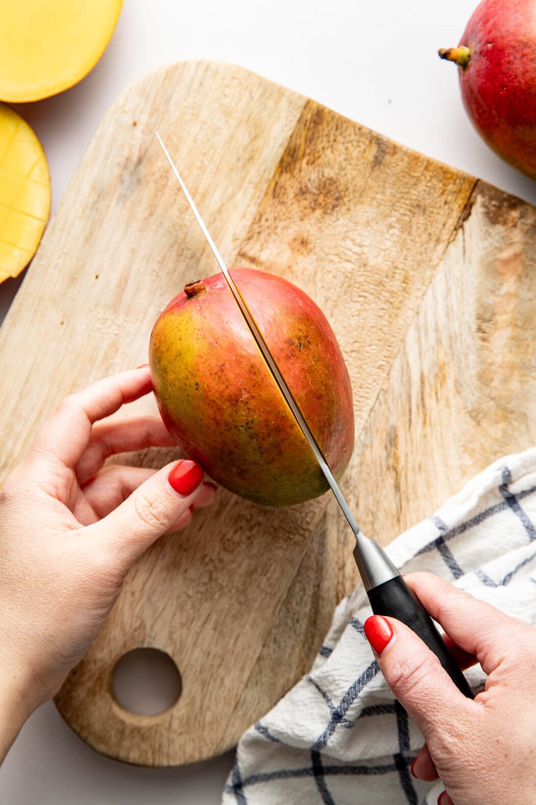 Overhead view of a hand holding a knife cutting into a mango on a wooden cutting board.