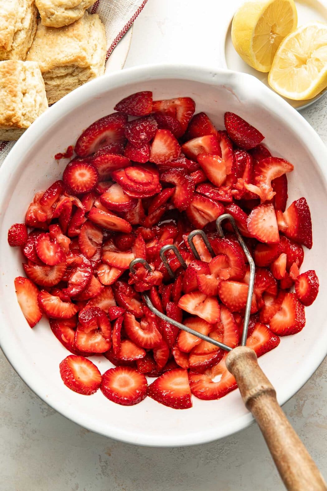 Overhead view of a bowl of sliced strawberries and a masher on the side of the bowl.