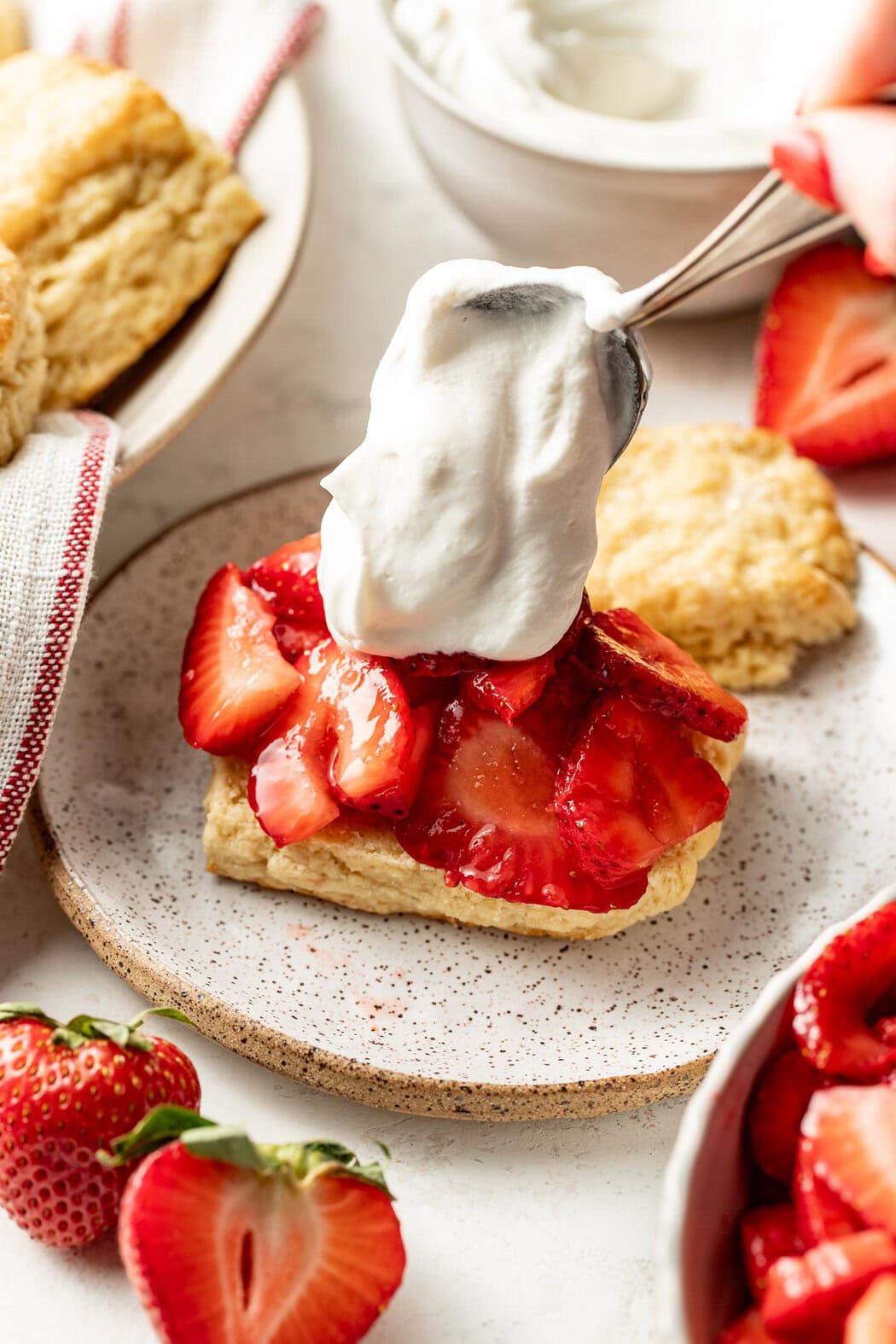 Overhead view of a spoon putting a dollop of whipped cream on top of a strawberry topped biscuit.