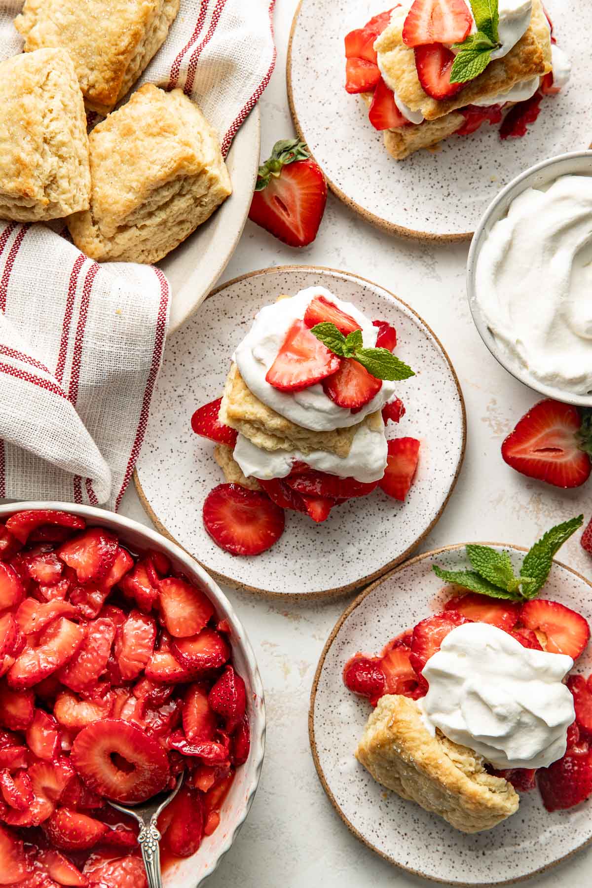 Overhead view of a tablescape of Strawberry Shortcake on plates, a bowl of biscuits, and a bowl of whipped cream all on a marbled countertop.