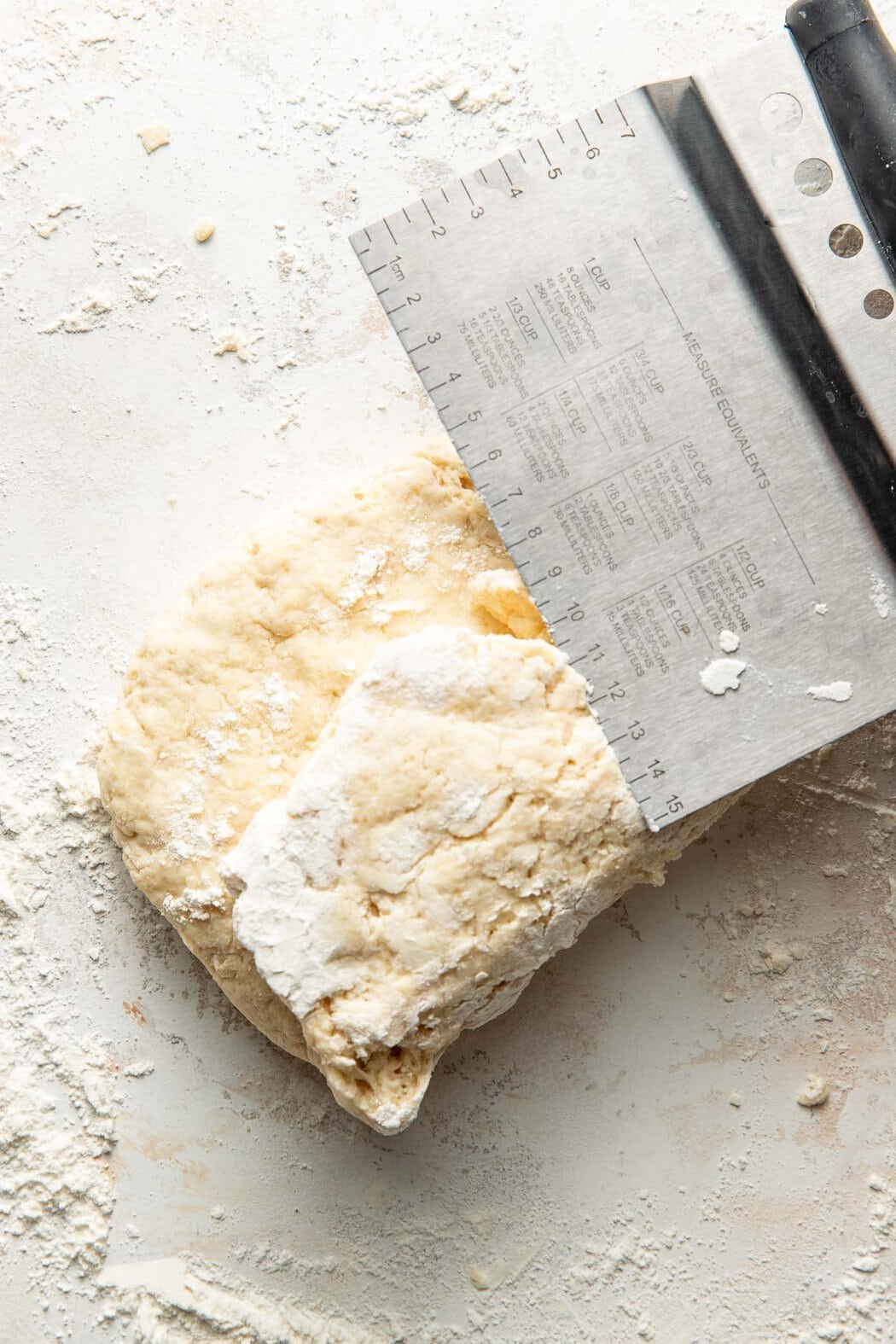 Overhead view of a piece of sweet cream biscuit dough on a floured countertop with a bench scraper on top.