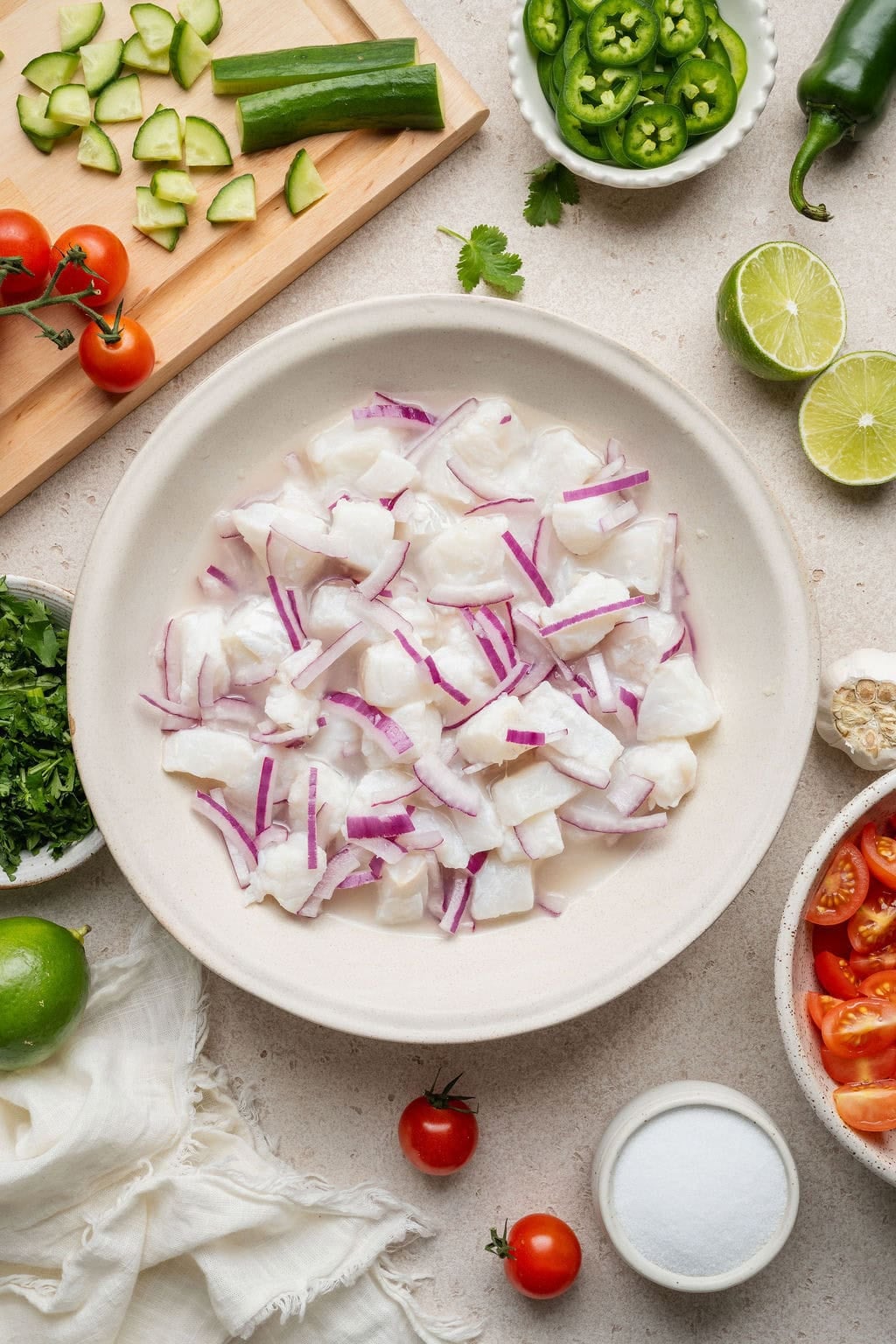 Overhead view of a bowl of white fish pieces and sliced red onion surrounded by fresh Ceviche ingredients. 