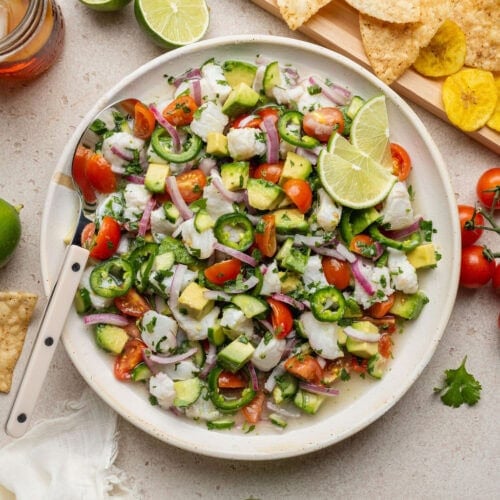 Overhead view of a bowl of fresh colorful ceviche topped with diced avocado and cilantro garnished with limes.