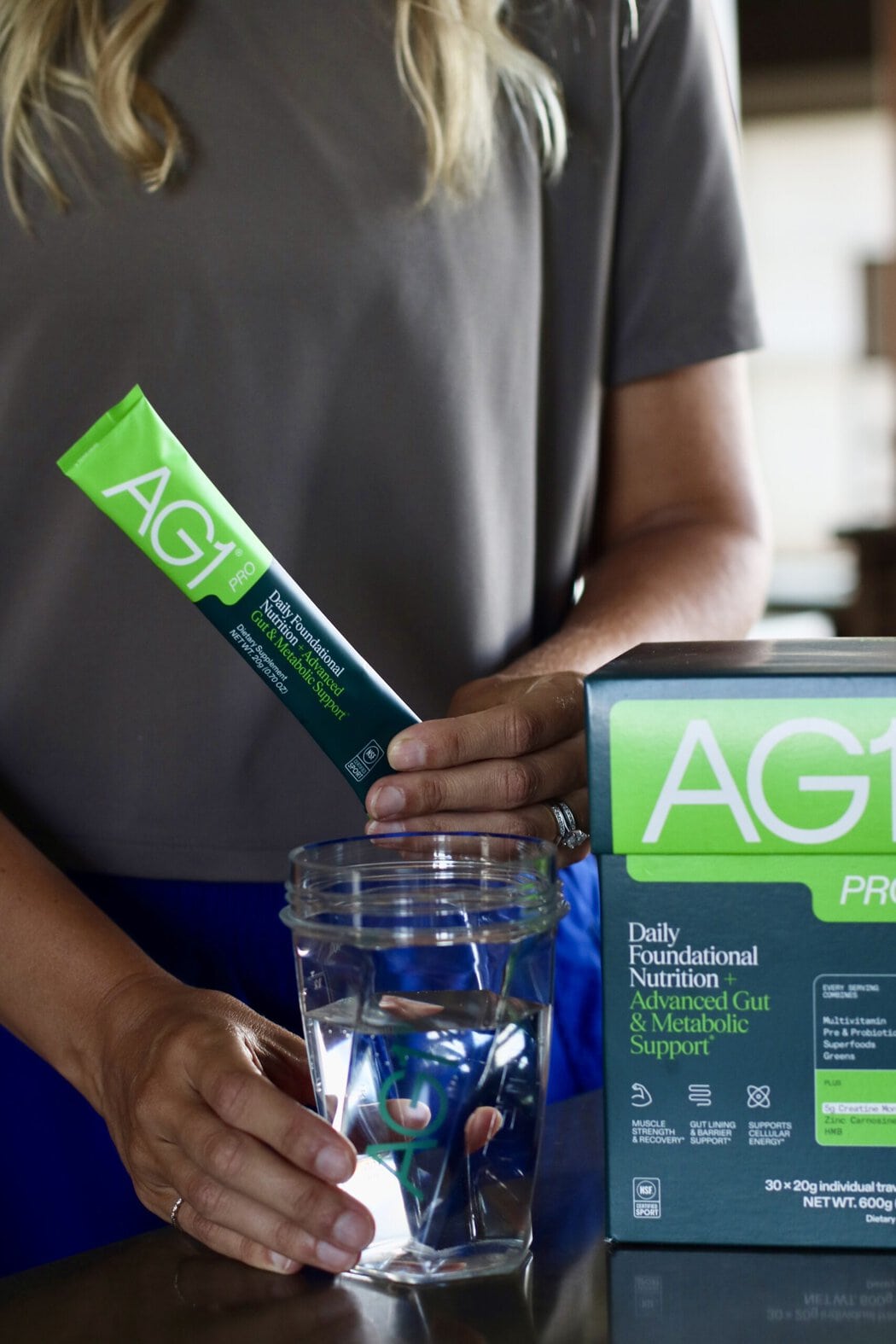 A woman holding an AG1 Pro packet in one hand and a glass of water in the other, a box of AG1 Pro next to her.