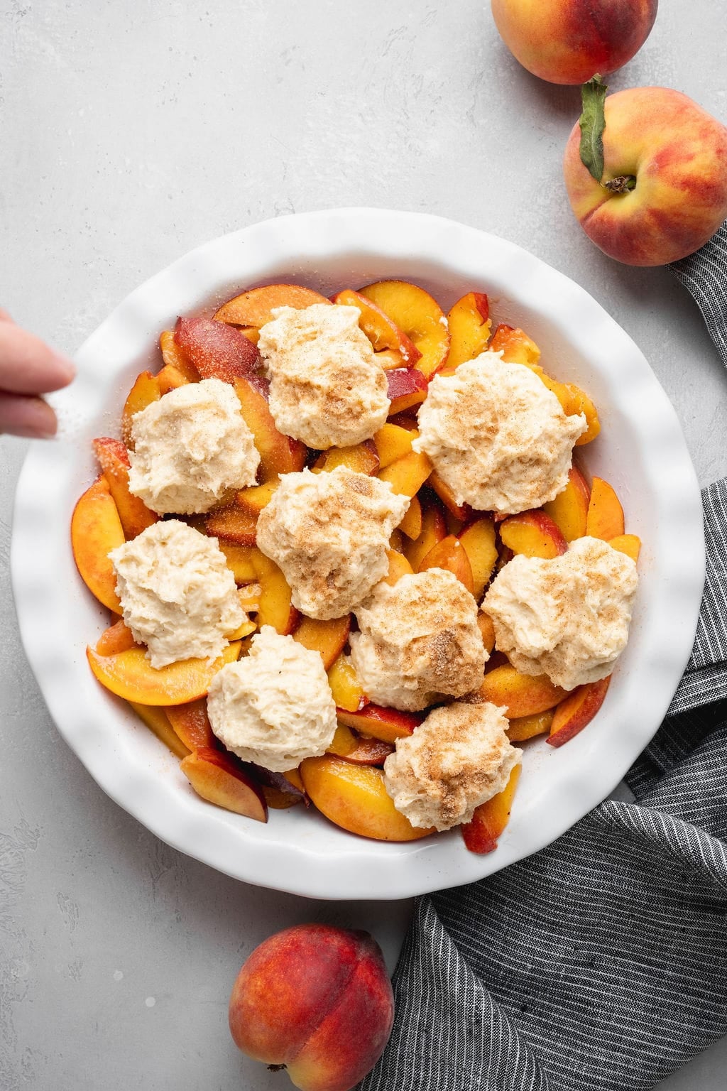 Peaches in the bottom of a round dish, dollops of cobbler dough on top, and a hand sprinkling sugar over the dough.