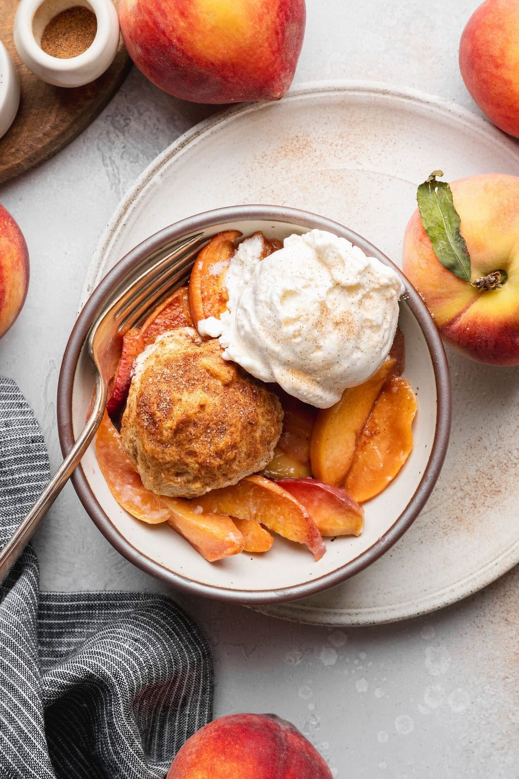 A bowl of peach cobbler topped with a biscuit and a scoop of vanilla ice cream, a fork on the side of the bowl.