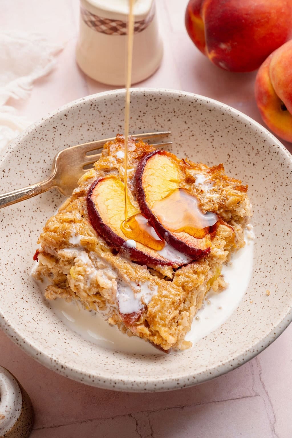 A serving of peach baked oatmeal in a bowl with a fork on the side, maple syrup being poured on top. 