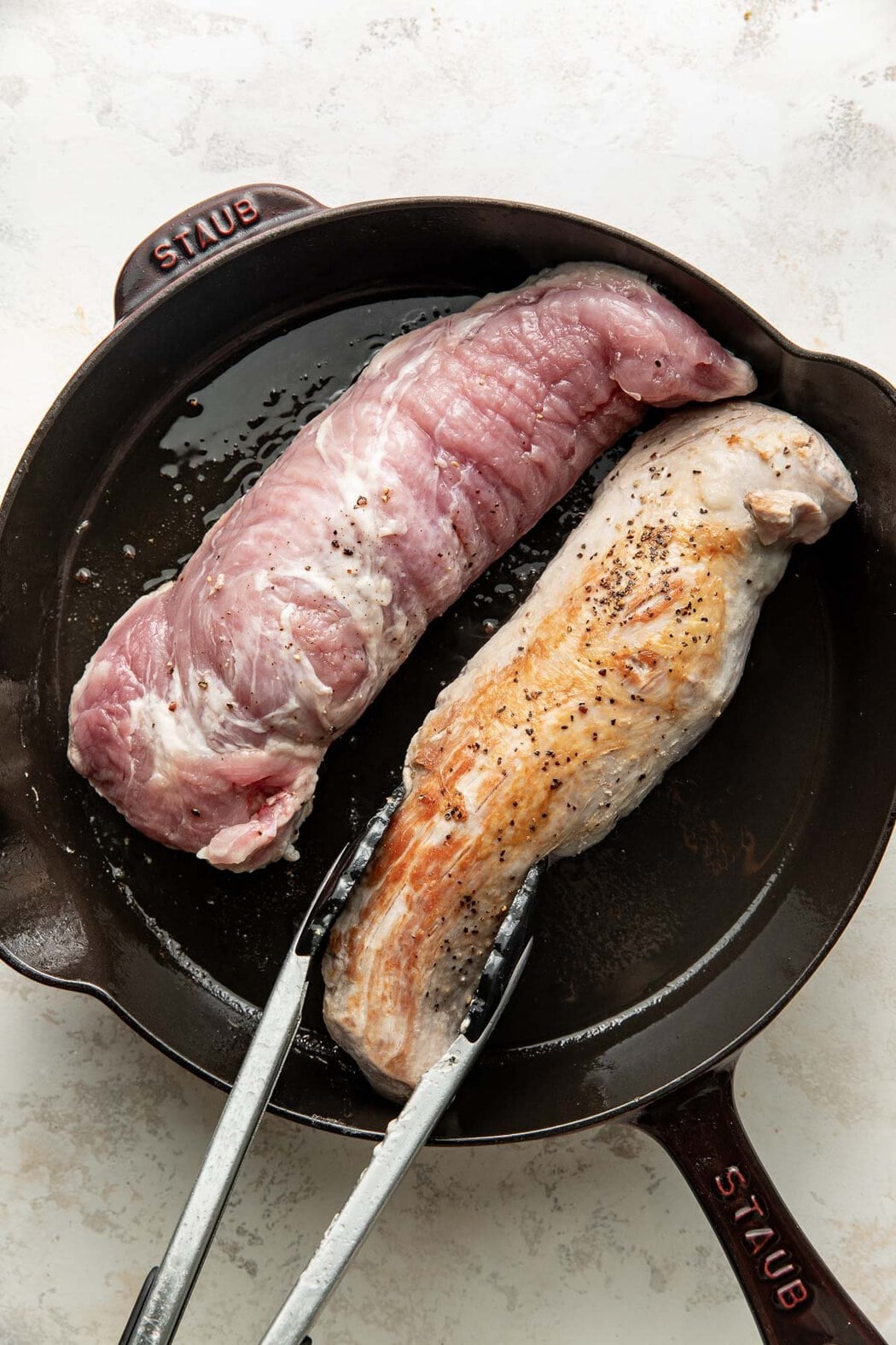 Two pork tenderloins being seared in a skillet, tongs flipping one over.