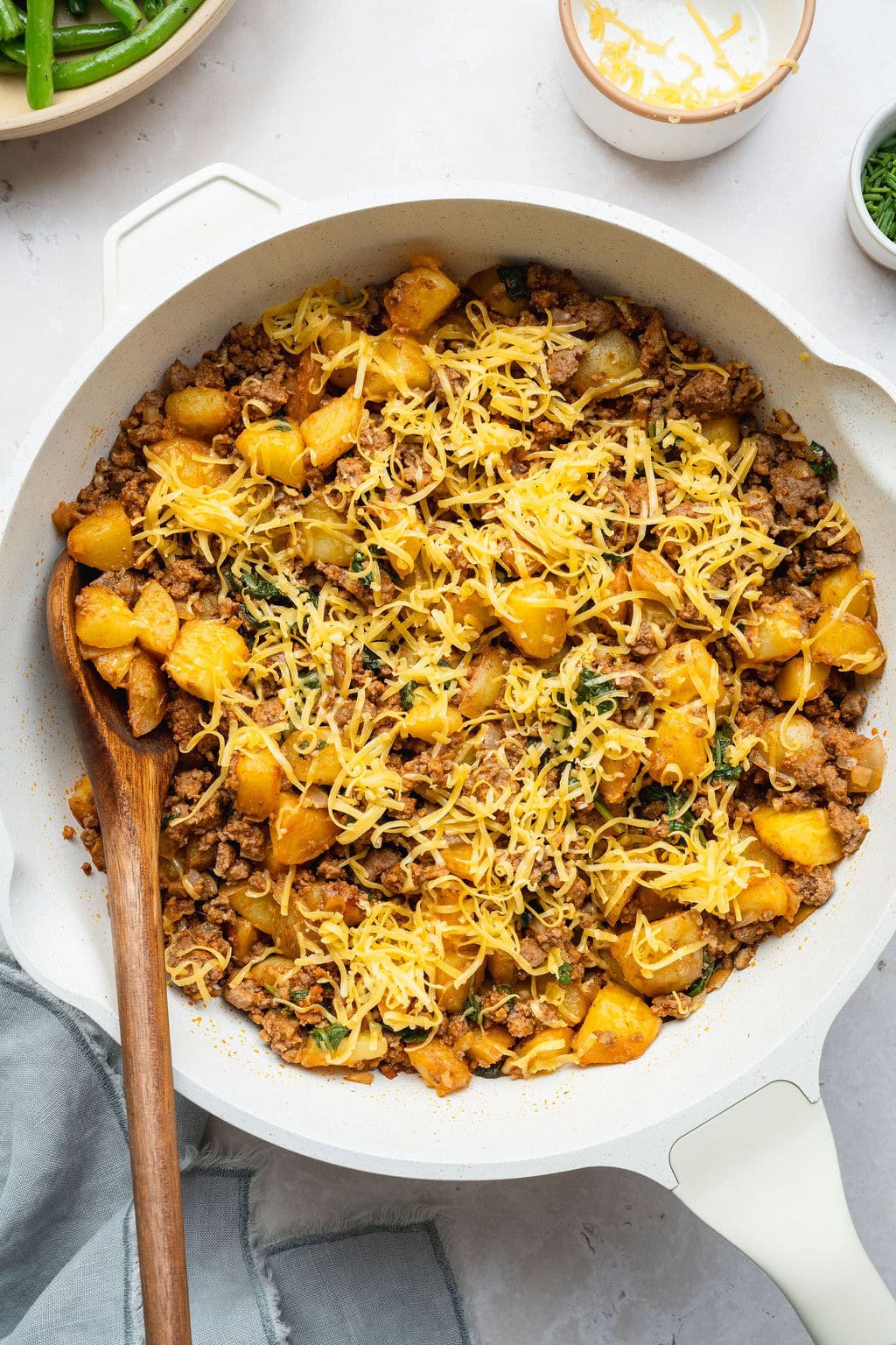 A ground beef and potatoes skillet topped with shredded cheese, a wooden spoon on the side.