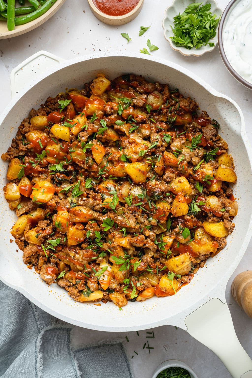 A cheesy ground beef and potatoes skillet topped with fresh herbs and ketchup.