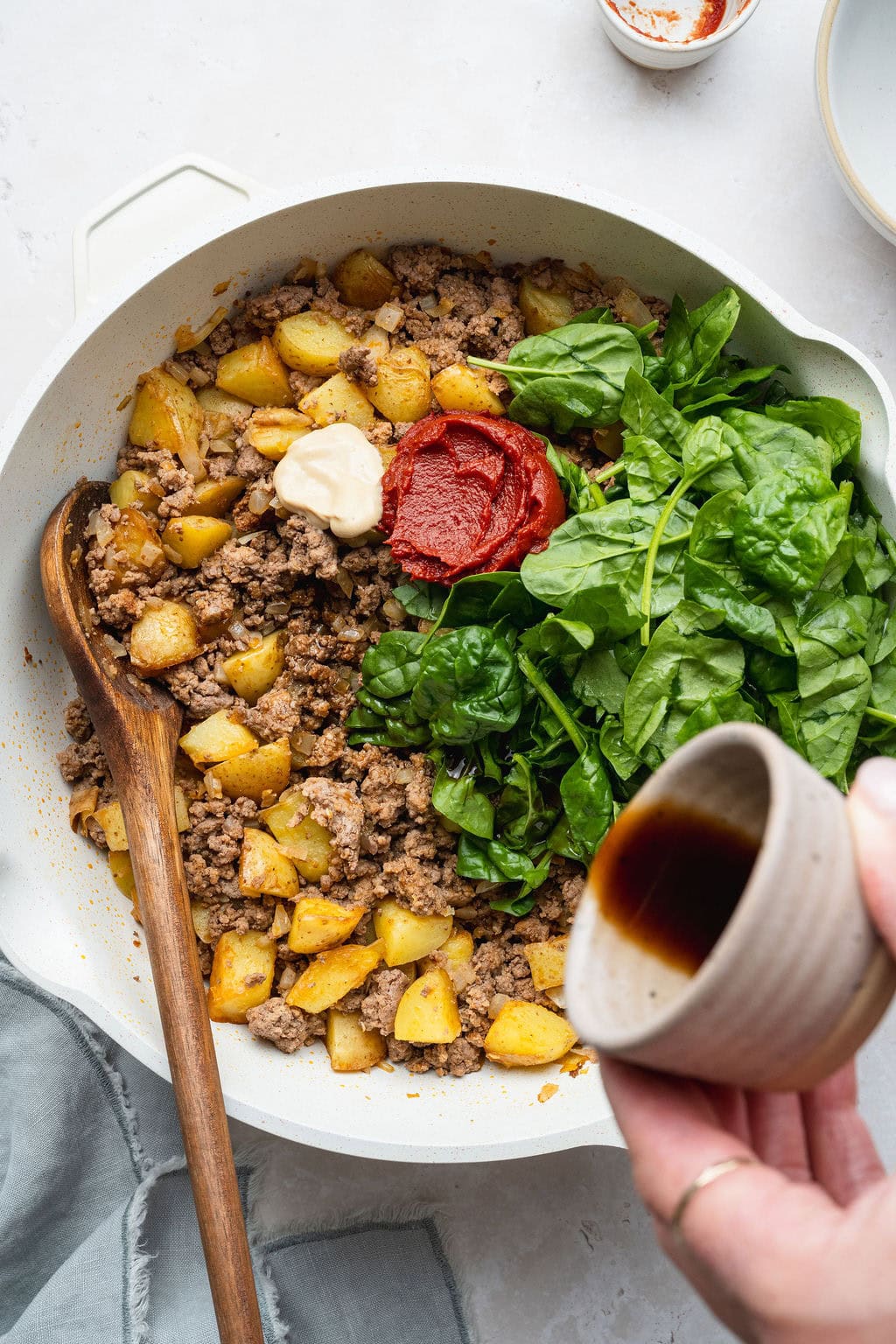 A skillet of browned ground beef, potatoes, spinach, tomato paste, and Dijon, Worcestershire sauce being poured on top.