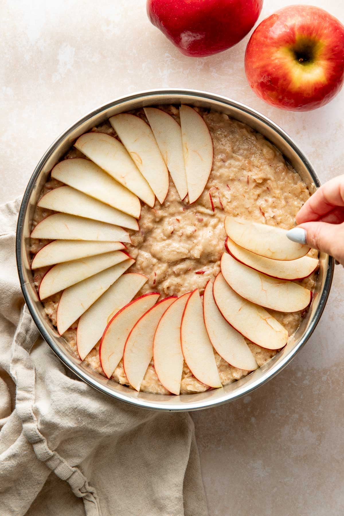 A woman's hand arranging apple slices over raw cake batter in a round pan.