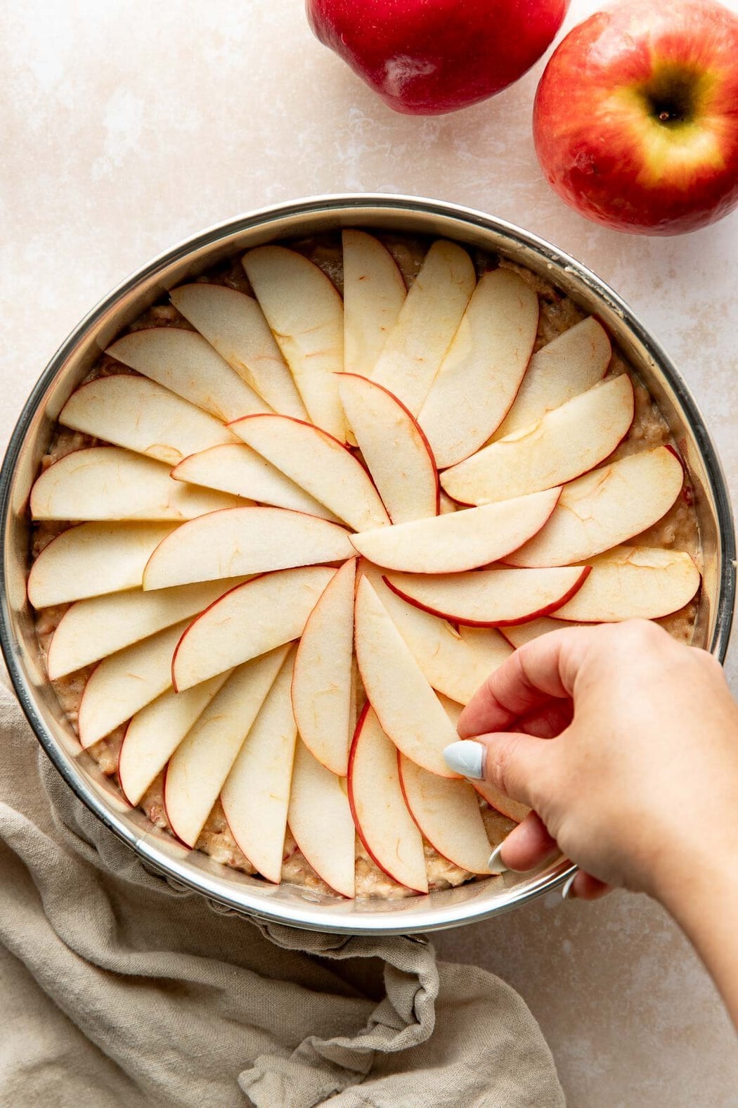 A woman's hand arranging apple slices in a circular pattern on top of raw apple cake batter in a round pan.