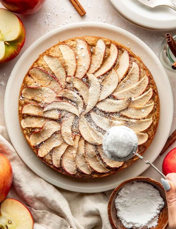 A round apple cake with slices of apples baked into the top on a platter with powdered sugar being sprinkled over top