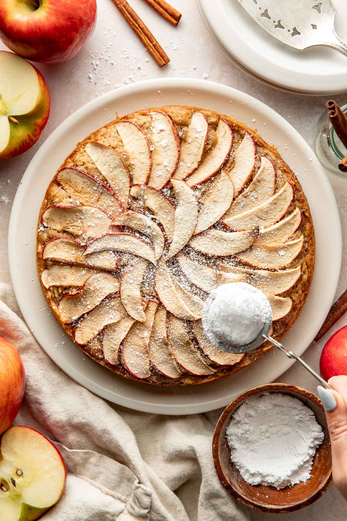 A woman's hand sprinkling powdered sugar over an apple cake on a plate.