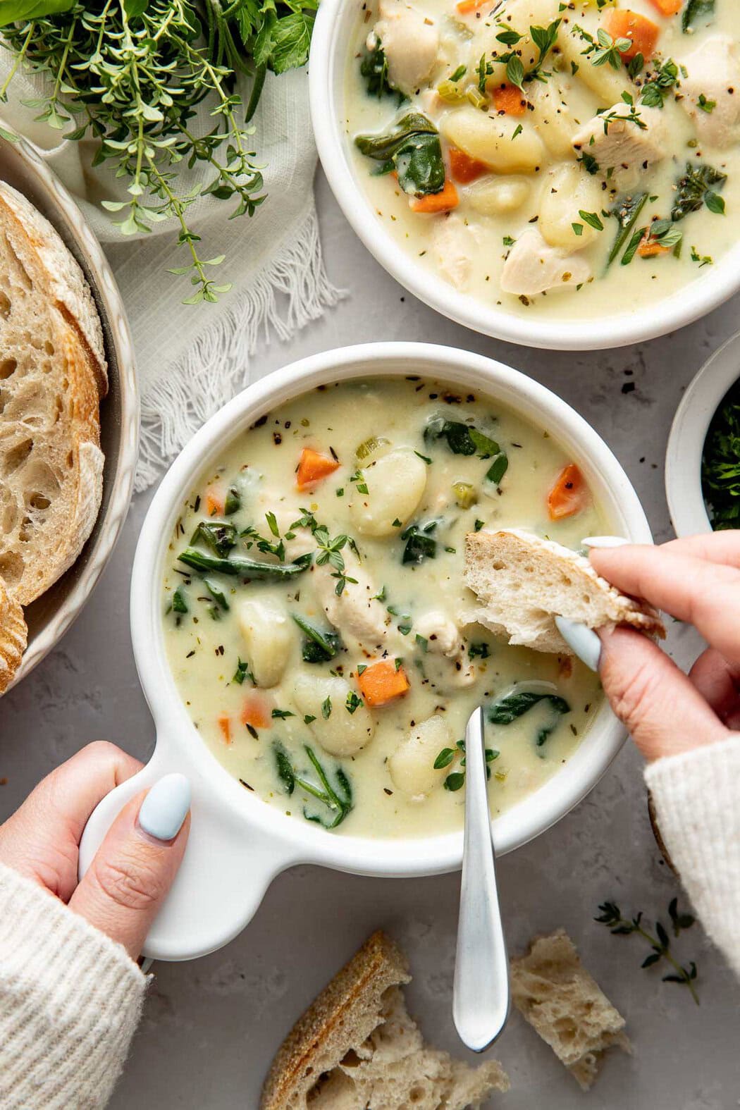A woman's hand holding a bowl of creamy chicken gnocchi soup, dipping a piece of bread into it.