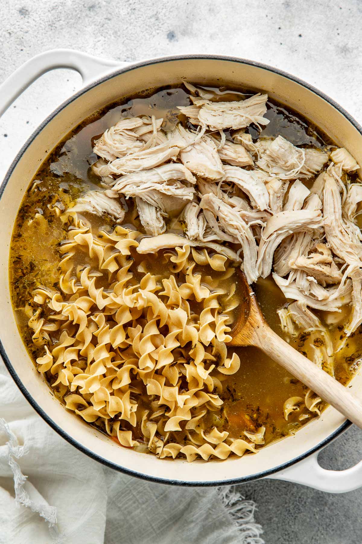 Shredded chicken and noodles being stirred into a pot of soup with a wooden spoon.