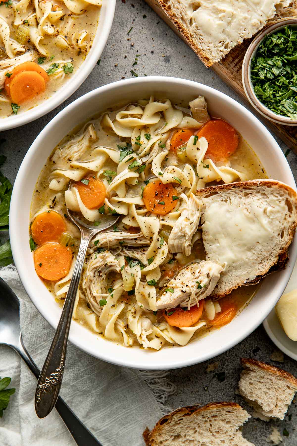 A bowl of homemade chicken noodle soup with a spoon sticking out and a buttered piece of bread.