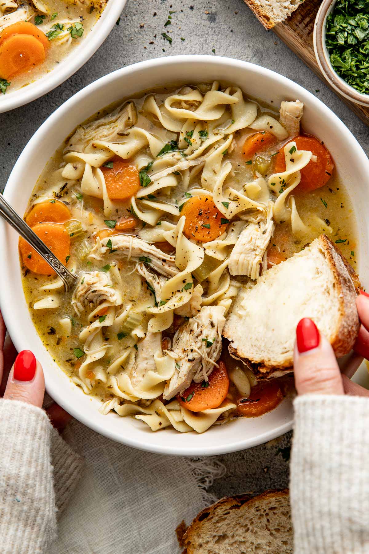 A bowl of chicken noodle soup, a spoon sticking out, and a woman's hand dipping a piece of buttered bread into the broth.