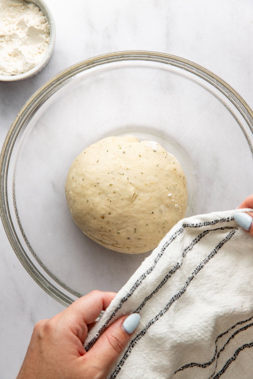 A ball of homemade pizza dough in a glass bowl being covered with a kitchen towel.