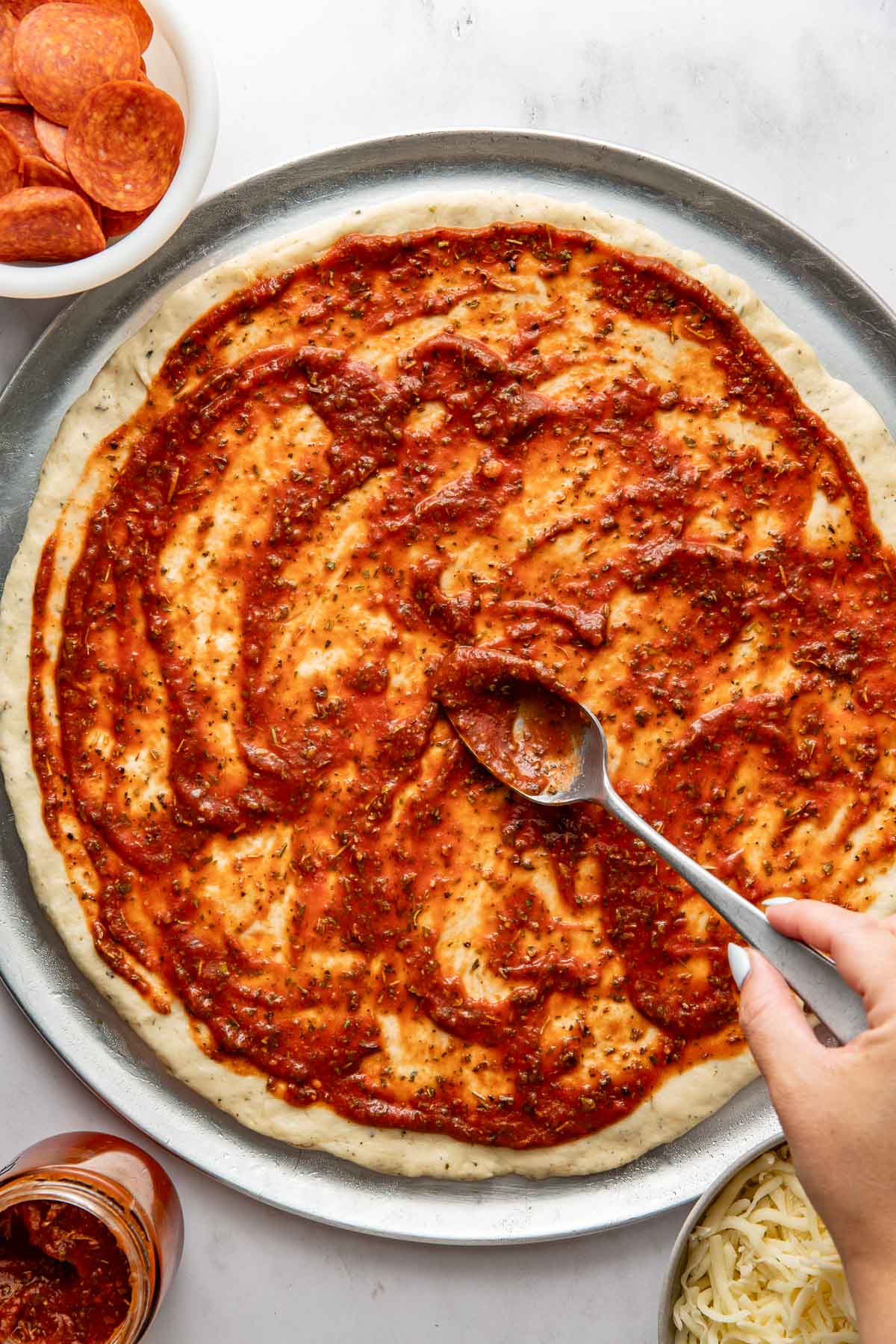 A hand spreading pizza sauce over a raw homemade pizza crust.