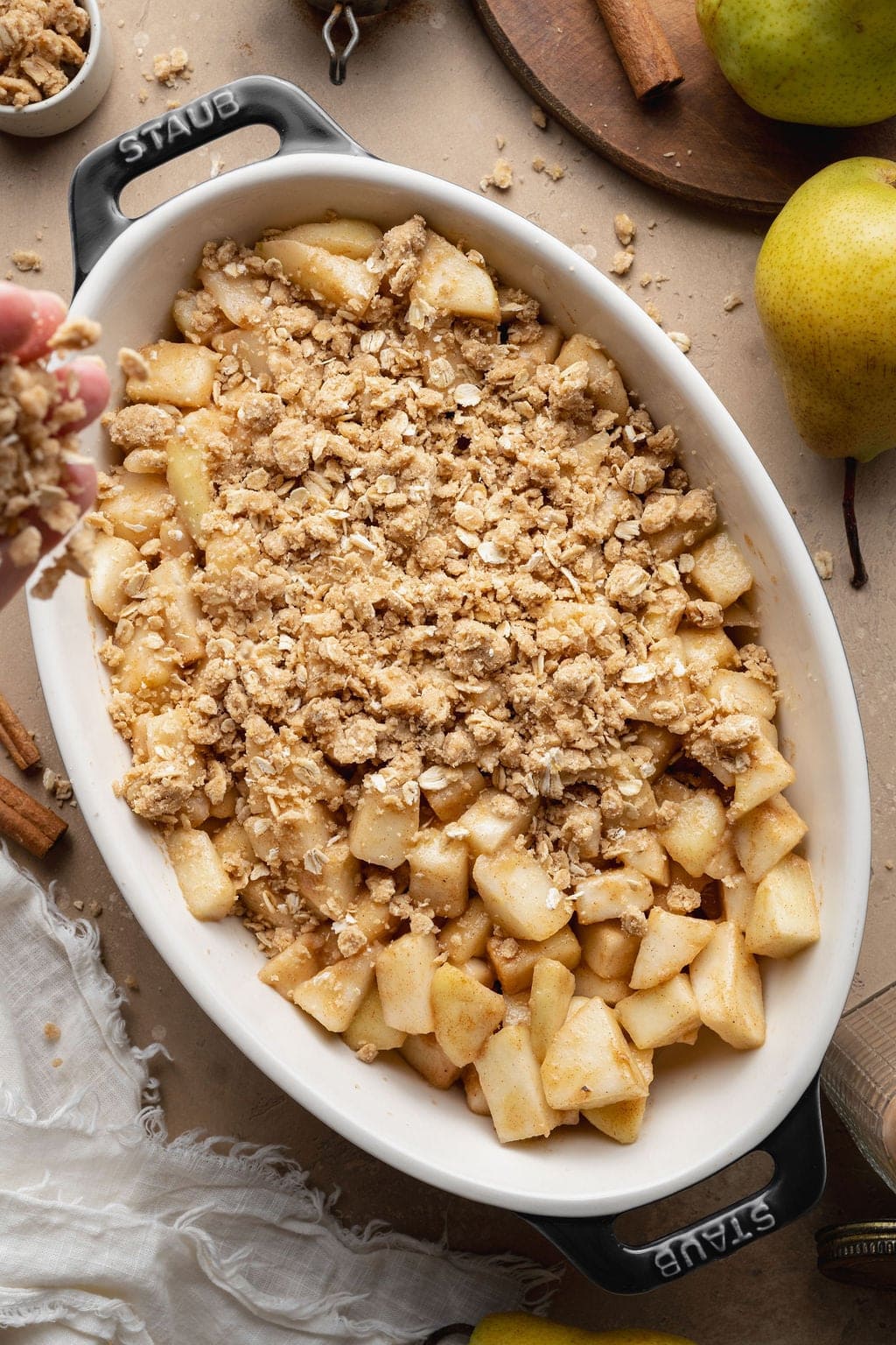 Crisp topping being sprinkled over spiced pear pieces in an oval baking dish.