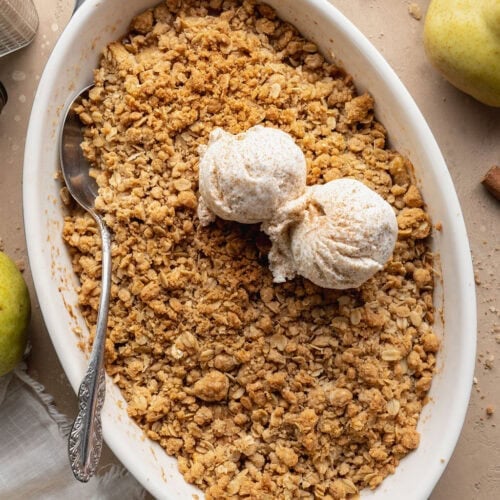 A pear crisp in an oval baking dish topped with two scoops of vanilla ice cream, a spoon on the side.