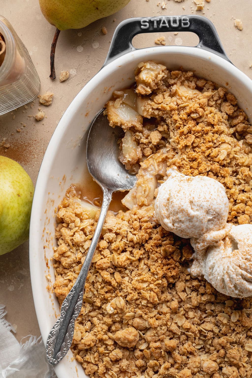 Pear crisp topped with two scoops of vanilla ice cream in an oval baking dish, a portion missing and a spoon sticking out.