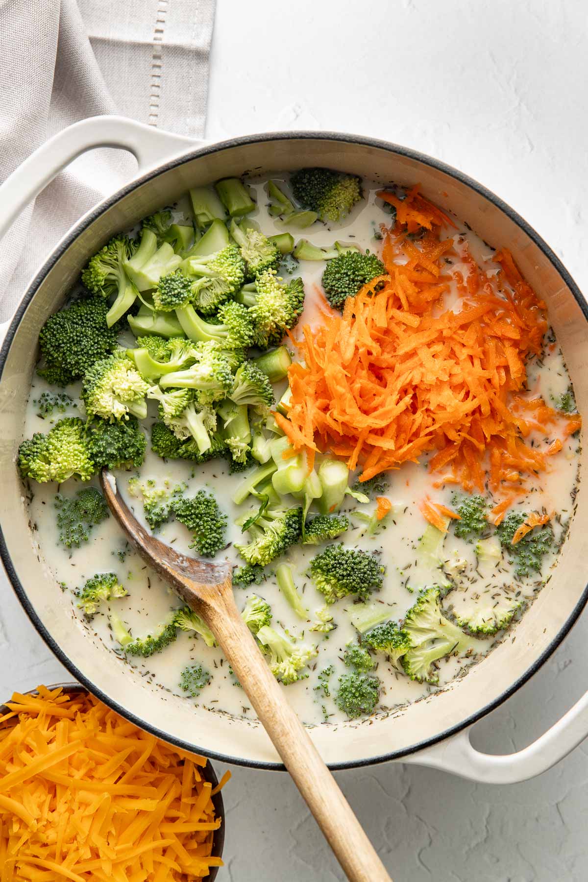 Shredded carrots and broccoli florets being stirred into a pot of soup with a wooden spoon.