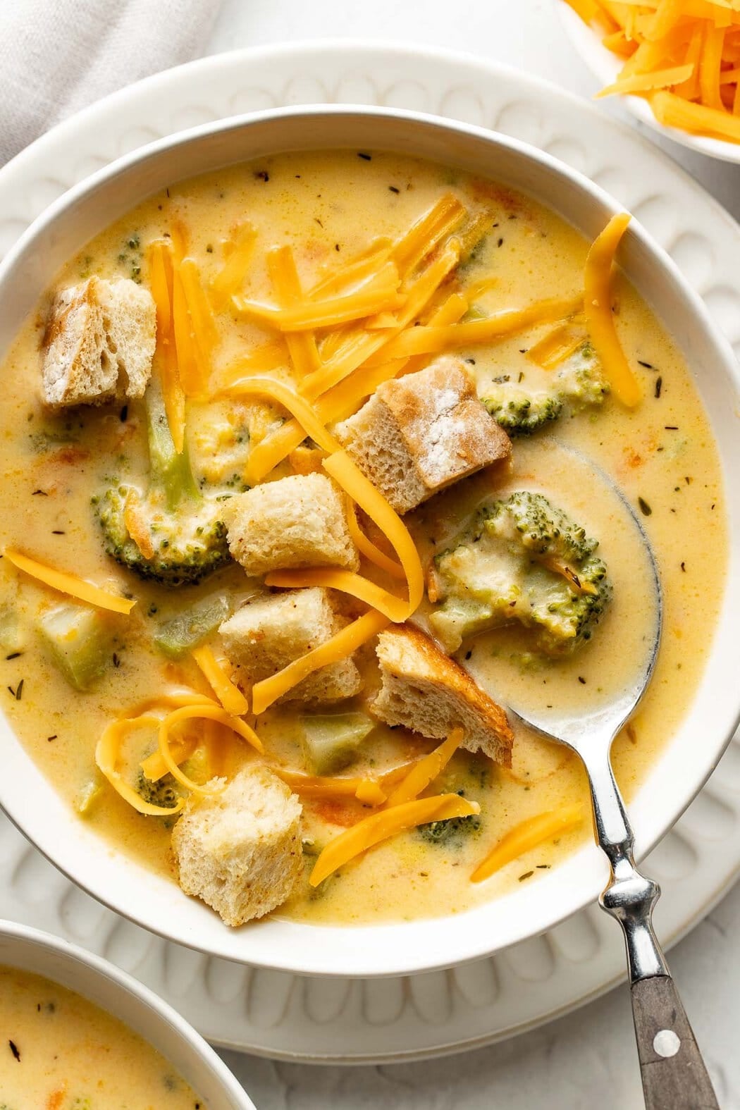 A bowl of broccoli cheddar soup topped with crusty bread pieces and shredded cheese, a spoon on top.