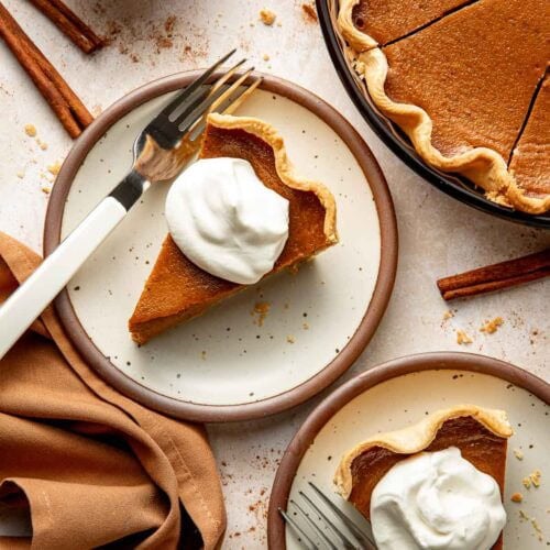 Two slices of pumpkin pie on separate plates topped with whipped cream, forks on the side.