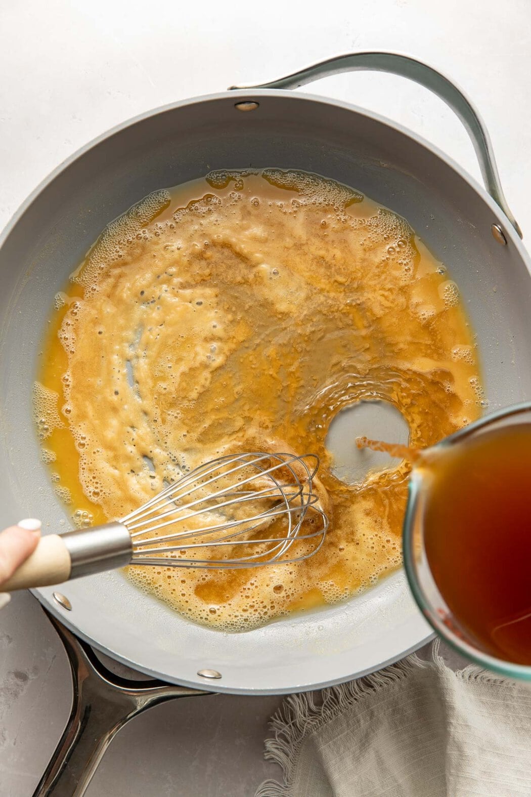 Broth being poured into a skillet, a hand whisking the mixture.