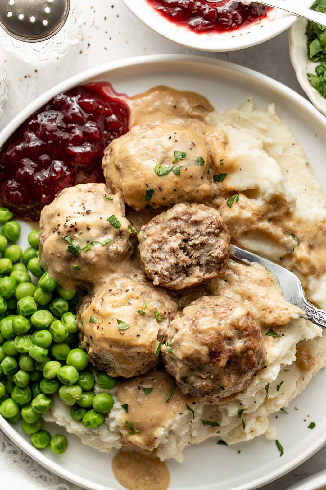 A plate of Swedish meatballs served over mashed potatoes with a side of peas and cranberry sauce, a fork holding a meatball missing a bite on top.