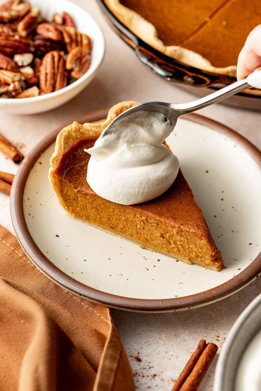 A woman's hand spooning a dollop of homemade whipped cream onto a piece of pumpkin pie.