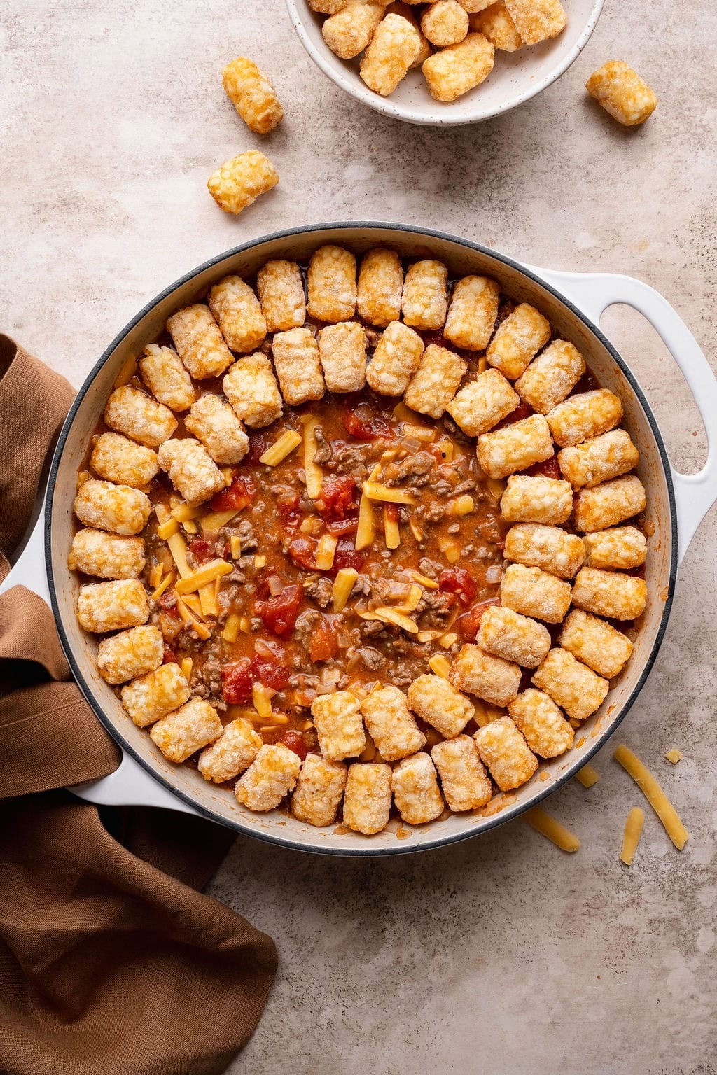 Frozen tater tots being arranged in a circular patter on top of a pot of cheeseburger casserole.
