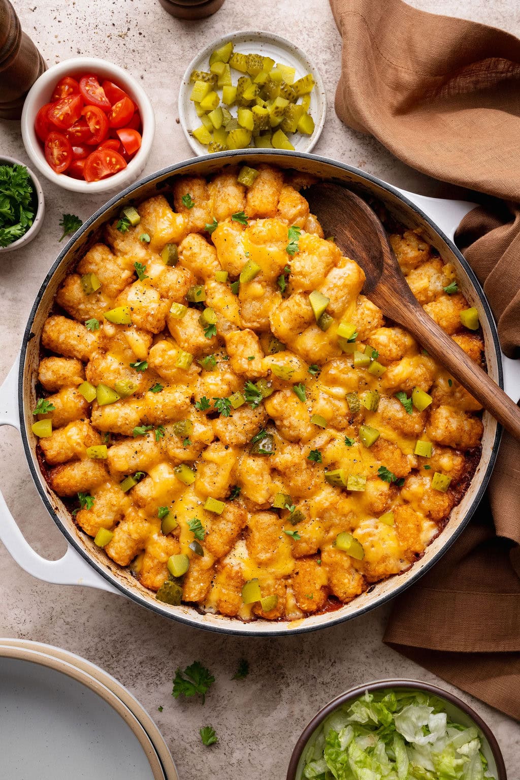 A pot of cheeseburger casserole topped with fresh herbs and pickles, a wooden spoon on top.