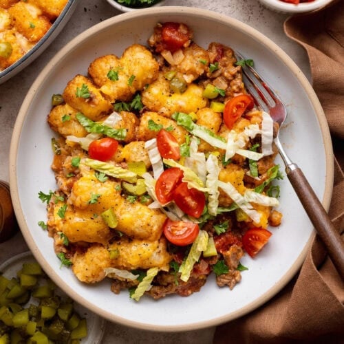 A serving of cheeseburger casserole in a bowl topped with shredded lettuce and tomatoes, a fork on the side.
