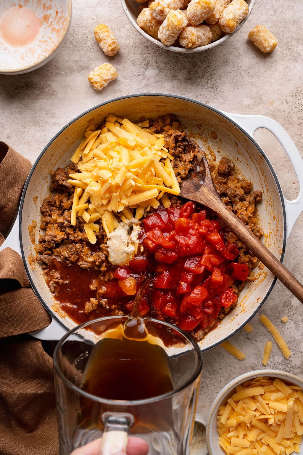 A pot of cooked ground beef and onions, shredded cheese, garlic, tomatoes, and broth being poured on top.